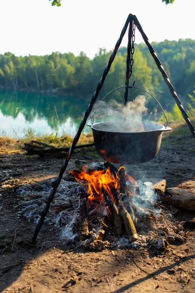 Seyahat ederken kazıkta yemek pişirmek. Ormanın yakınındaki bir gölün arka planında yanan melon şapkalı bir tripod. Kavramsal seyahat, yürüyüş ve macera.