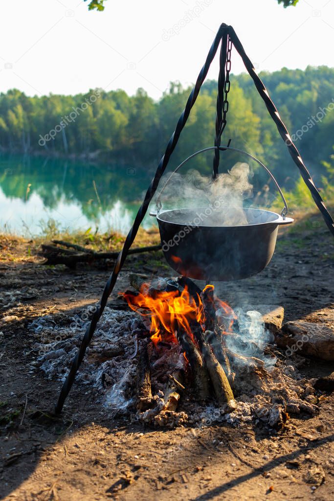 Cooking at the stake while traveling. Tripod with a bowler hat on a fire on the background of a lake near the forest. Conceptual travel, trekking and adventure.