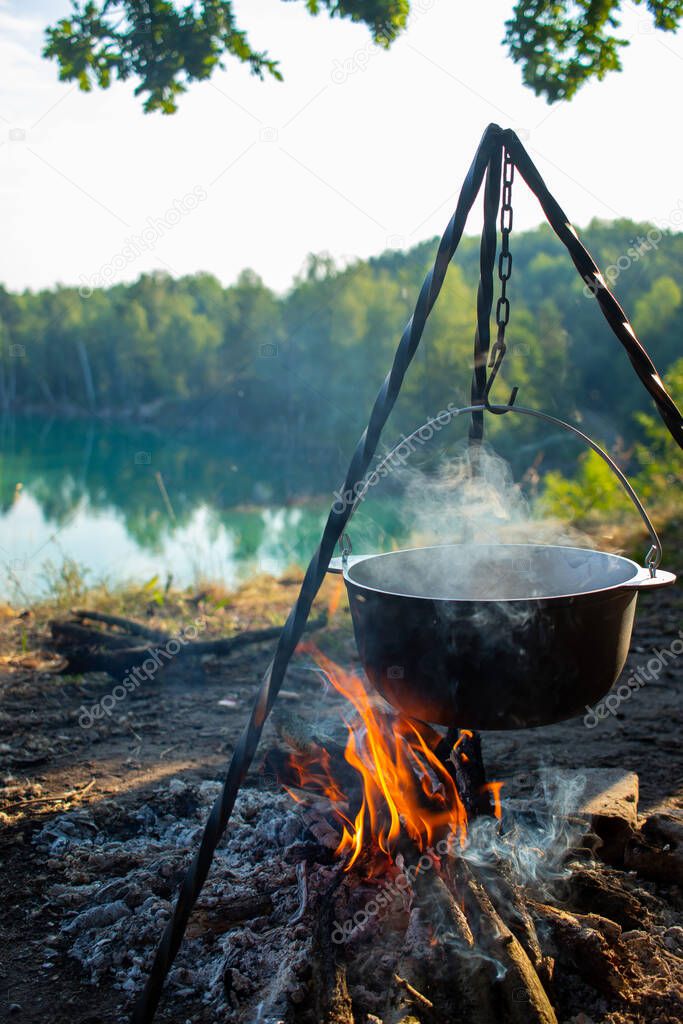 Cooking at the stake while traveling. Tripod with a bowler hat on a fire on the background of a lake near the forest. Conceptual travel, trekking and adventure. Field kitchen.