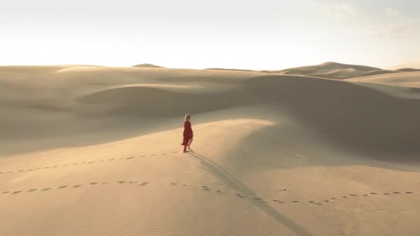 4K-Luftaufnahme eines Mädchens im flatternden roten Kleid, das bei Sonnenuntergang auf Sanddünen wandelt — Stockvideo