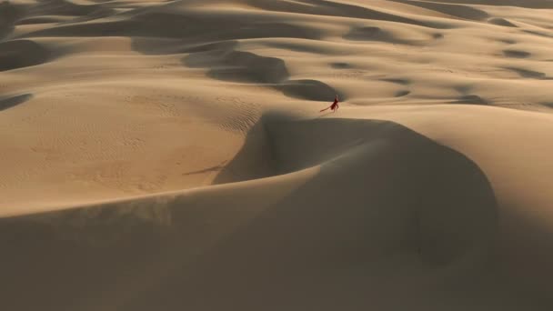 4K vue aérienne d'une femme solitaire marchant à travers les dunes de sable massives au coucher du soleil