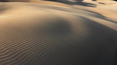 4K hava aracı görüntüsü altın gün batımında dalgalanan kum tepelerinin üzerinde uçuyor.