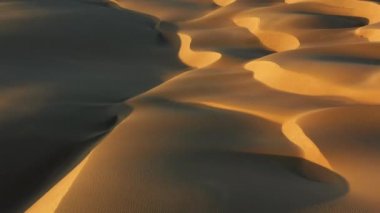 4K hava aracı görüntüsü altın gün batımında dalgalı kum tepelerinin üzerinde uçuyor.