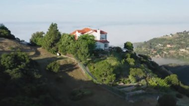 Yeşil dağın tepesinde ve güneş doğarken bulutların üzerinde evin manzaralı görüntüsü.