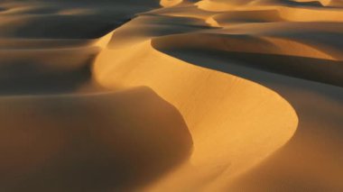 4K hava aracı görüntüsü altın gün batımında dalgalı kum tepelerinin üzerinde uçuyor.