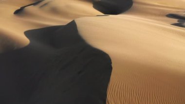 4K hava aracı görüntüsü altın gün batımında dalgalı kum tepelerinin üzerinde uçuyor.