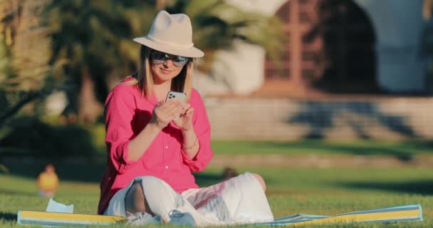 Розслаблена молода приваблива жінка за допомогою смартфона, що серфінгує соціальні медіа в парку — стокове відео