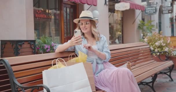 Hübsche Frau checkt Updates auf ihrem Smartphone, während sie auf einer Bank sitzt — Stockvideo