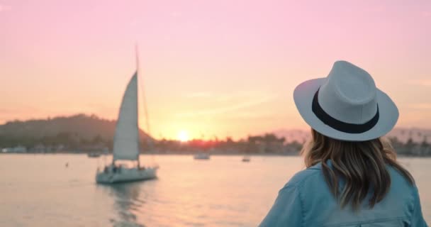 Elegantní dáma těší malebné růžové a zlaté západ slunce, zatímco plachetnice vstupuje do přístavu — Stock video