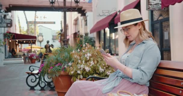 Mujer bonita está revisando el teléfono en verano, sentado en el banco en el casco antiguo cinematográfico — Vídeo de stock