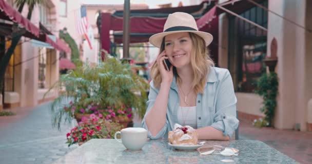 Позитивная улыбающаяся женщина разговаривает с другом по телефону сидя в уличном кафе — стоковое видео