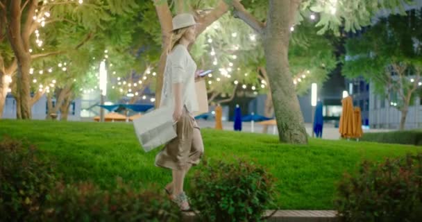 Mujer feliz en vestido elegante con bolsas de compras en la ciudad con iluminación nocturna — Vídeo de stock