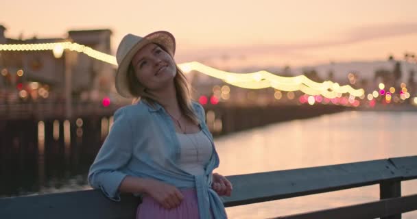 Elegante joven sonriente con sombrero disfrutando de la noche en el paseo marítimo en la ciudad costera — Vídeo de stock
