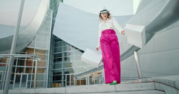 Mujer feliz en ropa de moda baila con bolsas de compras en el edificio moderno — Vídeos de Stock