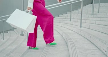 Pembe pantolonlu, beyaz alışveriş çantalı bir kadın modern şehrin merdivenlerinde yürüyor.