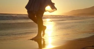 İnce kadın bacakları ve ayakları kumlu sahilde deniz dalgaları boyunca yürüyor.