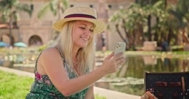 Yeşil parkta akıllı telefon kullanırken gülen mutlu sarışın kadın, 4K