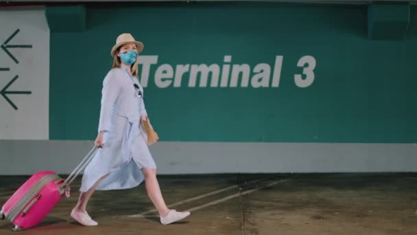 Стильная женщина в красочной маске для лица прогуливается по парковке аэропорта с чемоданом — стоковое видео