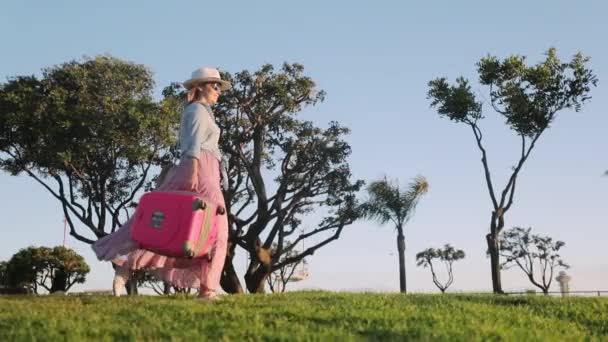 Fröhliche Frau begeistert von ihrer Reise und springt bei Sonnenuntergang mit rosa Reisetasche — Stockvideo