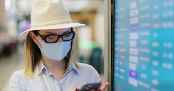 Женщина в маске проверяет свой телефон и расписание рейсов в пустом аэропорту, 4K — стоковое видео