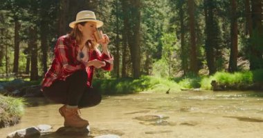 Ağır çekimde susamış bir kadın nehir kenarında oturmuş temiz su içiyor.