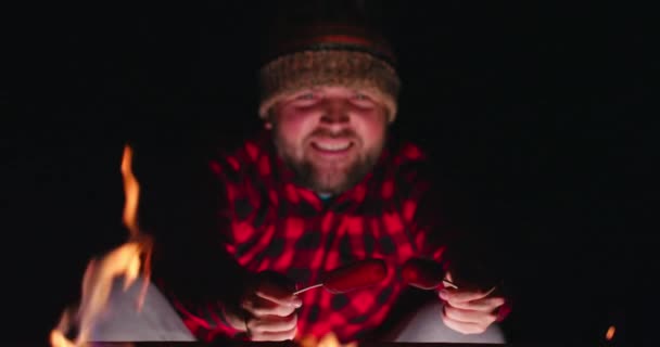 Omul zâmbitor prăjind cârnați pe foc de tabără și distrându-se făcând săruturi. Camping — Videoclip de stoc