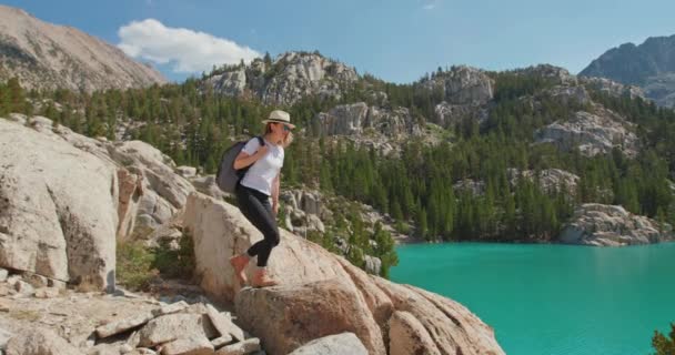 Женщина с поднятыми руками на вершине горы смотрит на живописное голубое озеро, 4K — стоковое видео