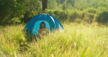Parlak yeşil çimenlikte, mavi kamp çadırında güneşli orman çayırında bir kadın.