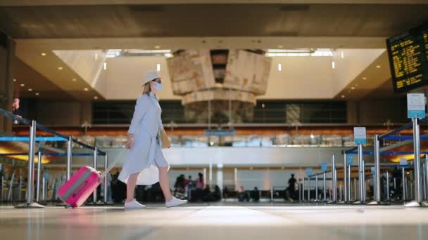 Zwolniony ruch stylowej kobiety w masce z torbą podróżną w pustym terminalu lotniska — Wideo stockowe