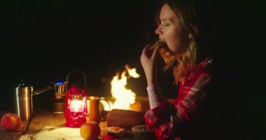 Kadın yemek ve içecekten zevk alıyor. Kamp alanında oturuyor ve arka planda yangın var.