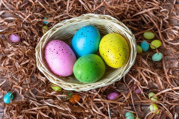 Four multicolor Easter eggs in a basket on wooden floor covered with straw and candies background. Easter celebratory wallpaper with copy space.