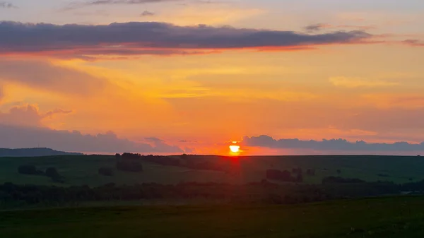 The sun sets on the horizon, the sky and the clouds are illuminated by the orange rays of the sun, summertime, the slopes of the hill are covered with green grass and trees.