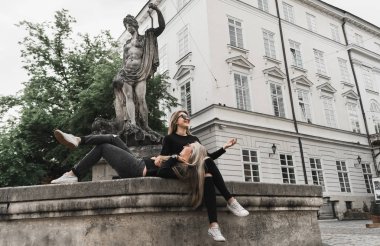 Fıskiyede oturan iki güzel kız. Fıskiyenin yanında siyah kıyafetler giyen arkadaşlar. Genç turistler eski şehirde konuşuyor ve gülüyor. Yaz tatillerinin tadını çıkaran pozitif bayanlar 