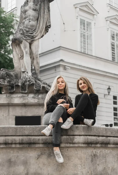 Two beautiful girls sitting at the fountain. Friends wearing in black clothes playing near fountain. Young tourists talking and laughing in the old city. Positive ladies enjoying summer vacations