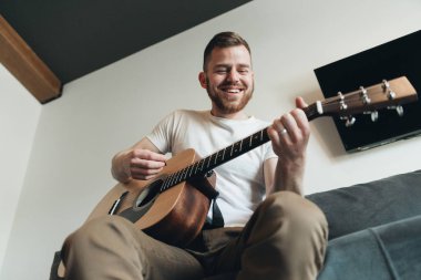 Klasik gitar çalan adam. Yakışıklı adam dairede müzik aleti ile rahatlatıcı. Akustik gitar çalan genç bir erkek. Müzisyen bir kanepede oturan ve gitar tutan