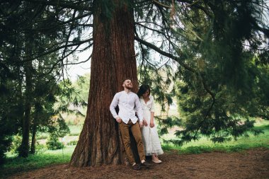 Neşeli genç evli çift el ele tutuşuyor ve ormanda yürüyor. Ağacın yanında poz veren tatlı çift. Güzel kız ve yakışıklı adam ormanda sıcak bir gün geçiriyor. Hafta sonu tatil konsepti