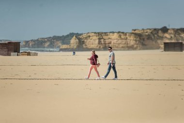 Mutlu genç çift plajda güneşli günlerin tadını çıkarıyor. Kumla oynayan kadın ve erkek. Okyanusa yakın bir aşk hikayesi. Gülümseyen çekici çifti kucaklayan ve binaların arka planda el ele tutuşan