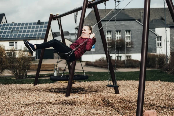 Young woman sitting on a swing in park. Pretty girl swinging on a swing against the urban landscape. Female feeling happy and healthy having fun in a sunny beautiful day