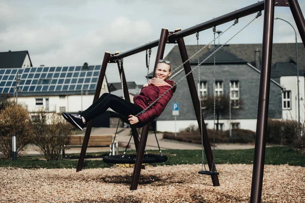 Young woman sitting on a swing in park. Pretty girl swinging on a swing against the urban landscape. Female feeling happy and healthy having fun in a sunny beautiful day