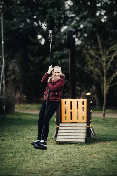 Young woman sitting on a swing in park. Pretty girl swinging on a swing against the urban landscape. Female feeling happy and healthy having fun in a sunny beautiful day