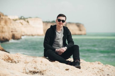 Uçurumun kenarında oturan genç turist. Kayalık sahildeki kayaların muhteşem manzarasının keyfini çıkaran adam. Okyanusa yakın siyah gözlüklü çekici çocuk. Dağ yaz manzara