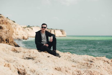 Uçurumun kenarında oturan genç turist. Kayalık sahildeki kayaların muhteşem manzarasının keyfini çıkaran adam. Okyanusa yakın siyah gözlüklü çekici çocuk. Dağ yaz manzara