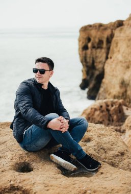 Uçurumun kenarında oturan genç turist. Kayalık sahildeki kayaların muhteşem manzarasının keyfini çıkaran adam. Okyanusa yakın siyah gözlüklü çekici çocuk. Dağ yaz manzara