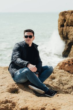 Uçurumun kenarında oturan genç turist. Kayalık sahildeki kayaların muhteşem manzarasının keyfini çıkaran adam. Okyanusa yakın siyah gözlüklü çekici çocuk. Dağ yaz manzara