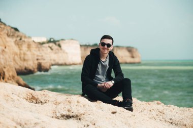 Uçurumun kenarında oturan genç turist. Kayalık sahildeki kayaların muhteşem manzarasının keyfini çıkaran adam. Okyanusa yakın siyah gözlüklü çekici çocuk. Dağ yaz manzara
