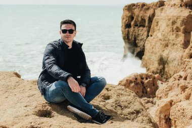 Uçurumun kenarında oturan genç turist. Kayalık sahildeki kayaların muhteşem manzarasının keyfini çıkaran adam. Okyanusa yakın siyah gözlüklü çekici çocuk. Dağ yaz manzara