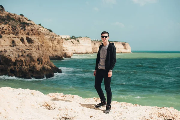 Uçurumun kenarında duran genç turist. Kayalık sahildeki kayaların muhteşem manzarasının keyfini çıkaran adam. Okyanusa yakın siyah gözlüklü çekici çocuk. Dağ yaz manzara