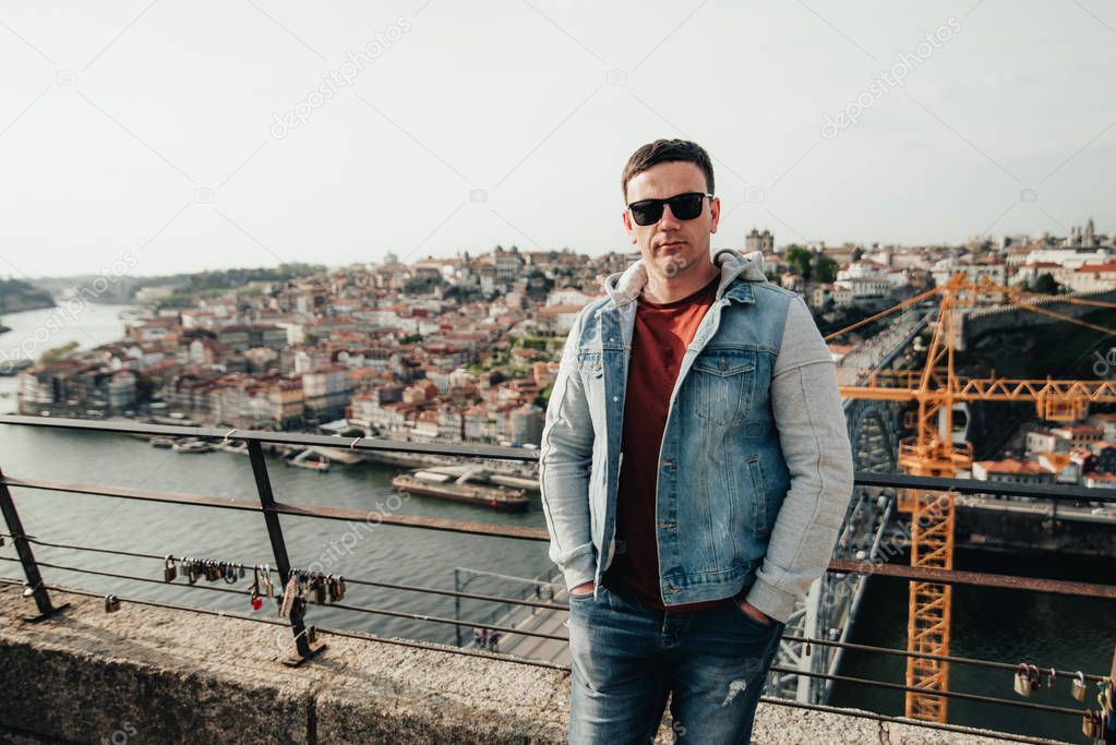 Handsome man staying on the bridge on the city background. Tourist is walking on the quay. The young guy admires the beauty of the old city in Portugal.