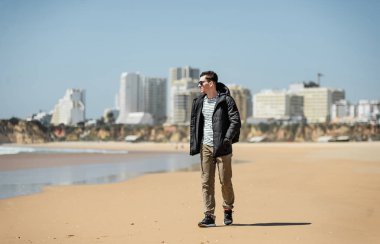 Genç adam sahilde güneşli günlerin tadını çıkarıyor. Portre güzel guyl yürüyüş ve kum ile oynarken. Okyanus kenarında poz veren gülümseyen bir erkek. Portekiz'de Tatil