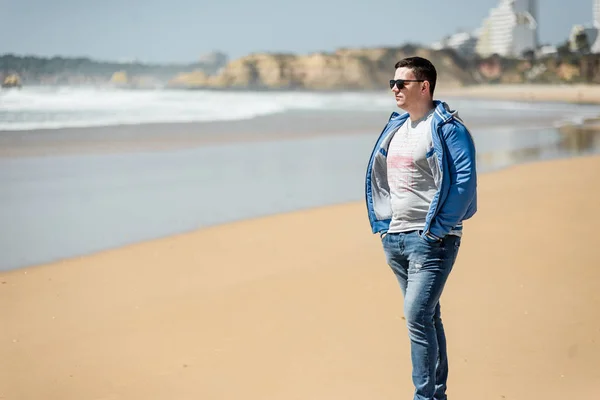 Genç adam sahilde güneşli günlerin tadını çıkarıyor. Portre güzel guyl yürüyüş ve kum ile oynarken. Okyanus kenarında poz veren gülümseyen bir erkek. Portekiz'de Tatil
